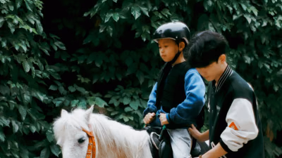 Kind mit Reithelm und Sicherheitsweste sitzt auf einem weißen Pony, ein Erwachsener führt und sichert es