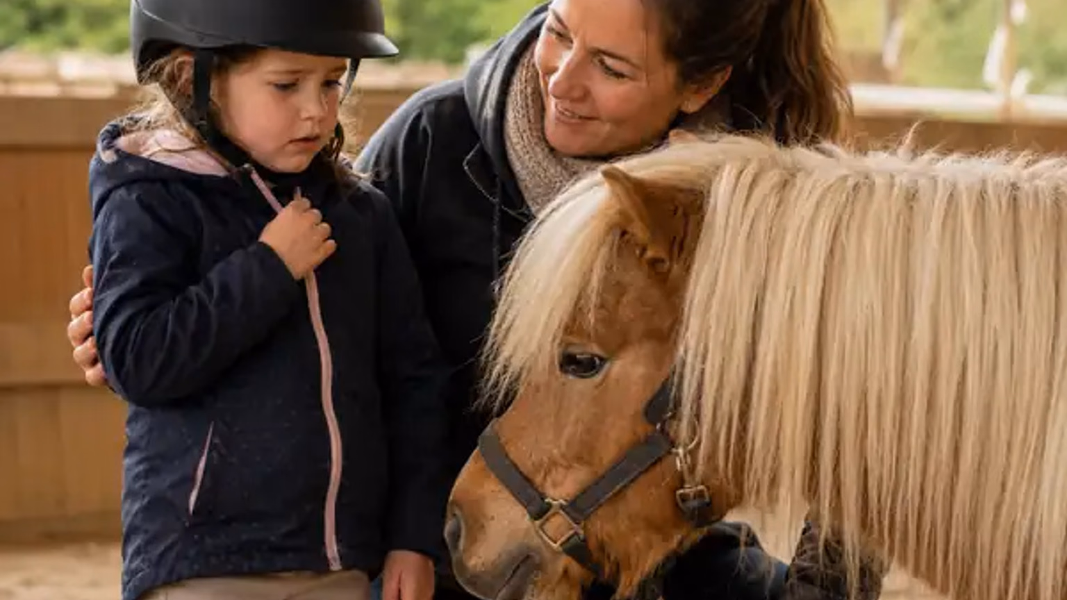 Vorsichtiges Kind berührt ein kleines Pony, während eine Erwachsene ruhig danebensteht