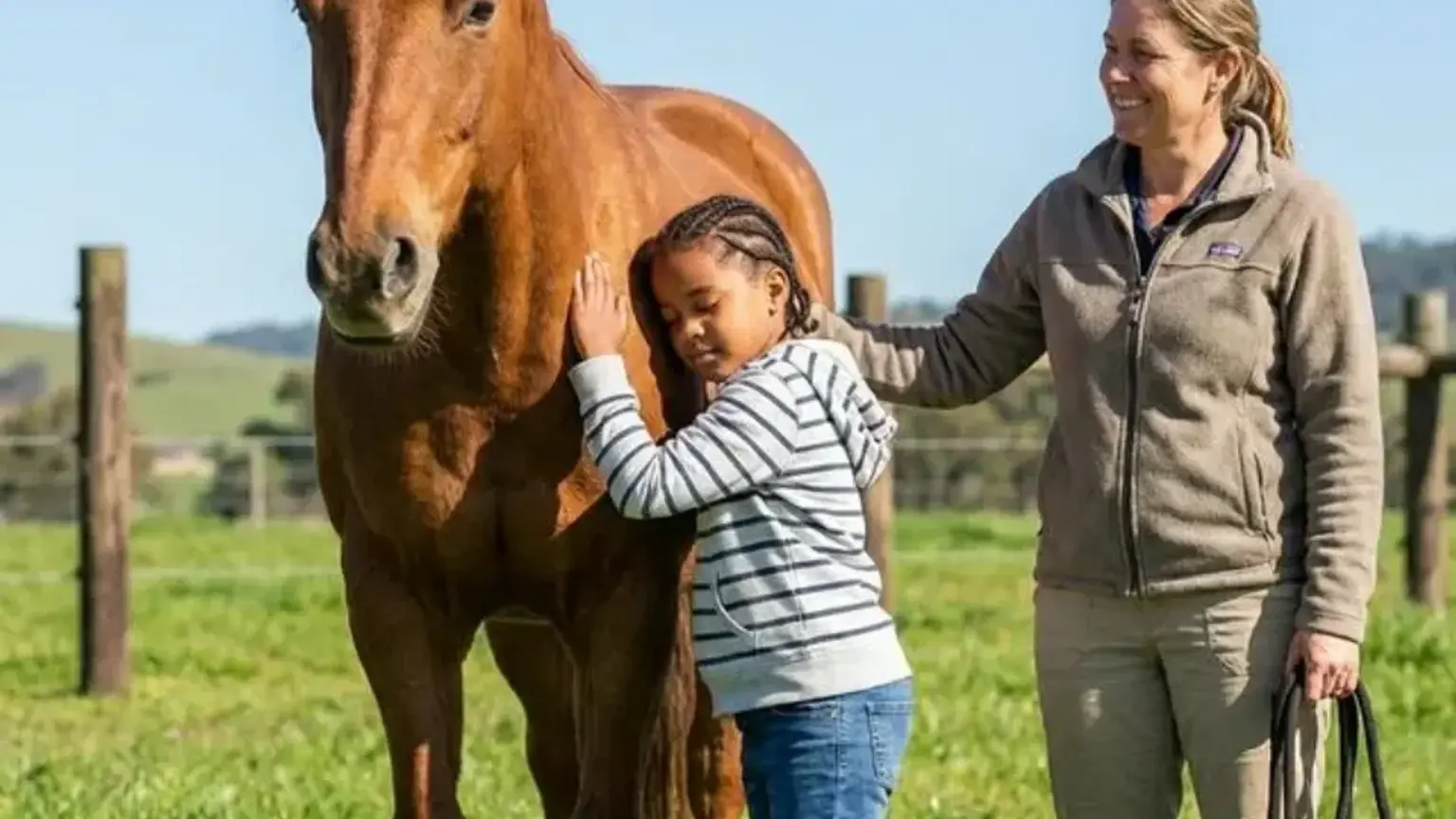Ein sonniger Tag auf der Koppel: Ein Kind streichelt das Pferd ruhig am Hals, während die Reittherapeutin lächelnd daneben steht